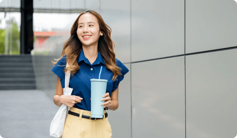 Asian woman with blue collared shirt walking outside a corporate building with a reusable water cup