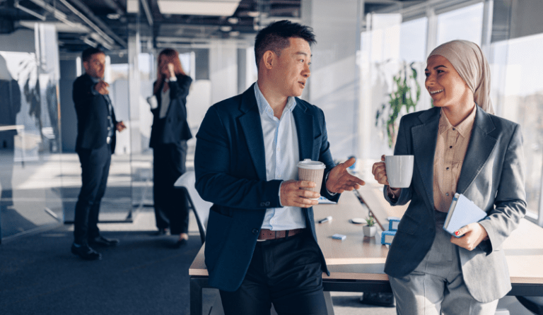 two co-workers at a desk talking and drinking coffee